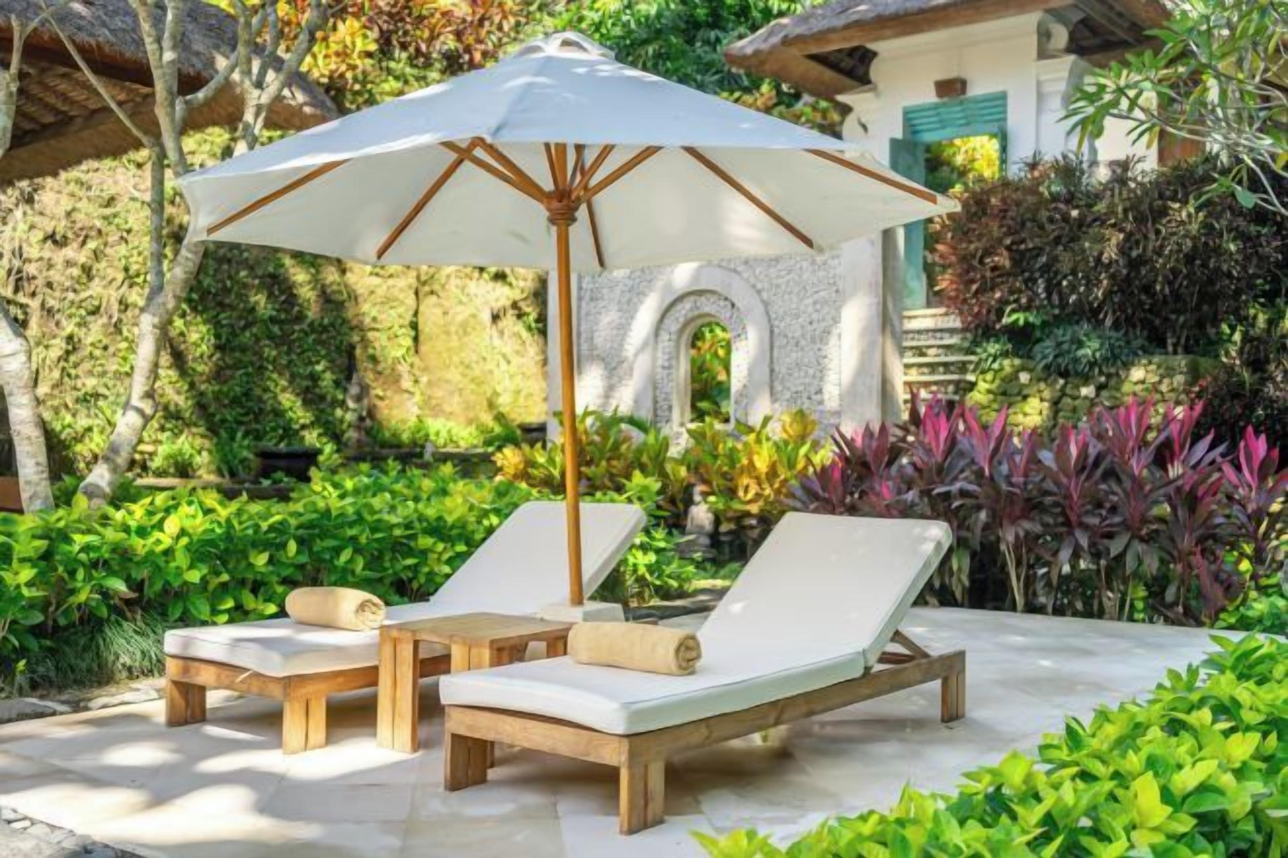 A patio with two lounge chairs and a large umbrella, surrounded by lush greenery and a stone wall.