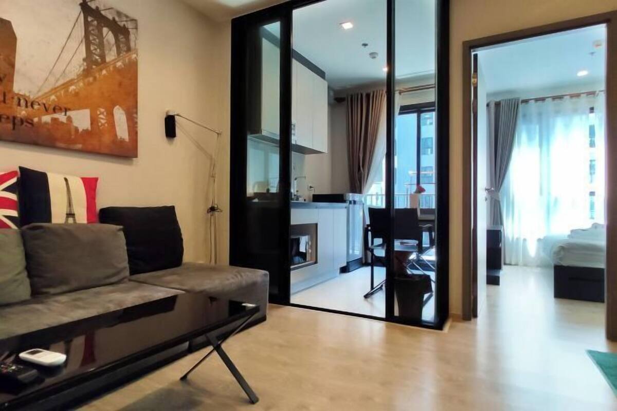 Un salon moderne avec un canapé, une table basse et un tableau représentant un pont. Une porte-fenêtre coulissante donne accès à un coin repas et à une cuisine.