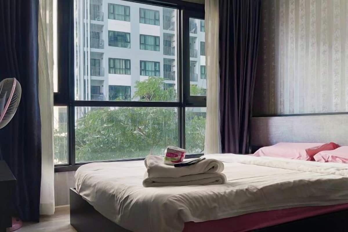 Une chambre d’hôtel avec une grande fenêtre, un lit avec une literie blanche, un ventilateur et un petit objet posé sur le lit.