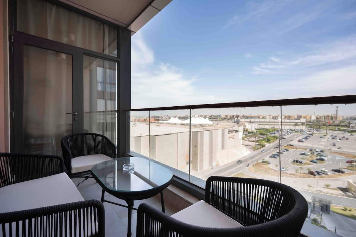 A balcony with a glass table and chairs, offering a view of a cityscape with buildings and a parking area.