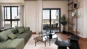 A modern living room with a green sofa, a glass coffee table, and a dining area with a small table and chairs. There is a large window with curtains, a potted plant, and a bookshelf against the wall. - Aeon Towers Residence - BirdNest (6th of October City)