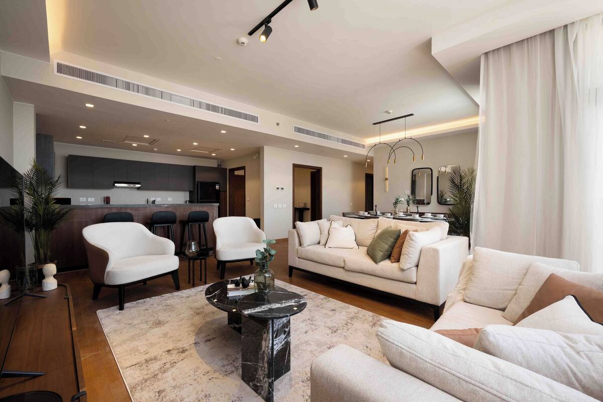 A modern living room with a sofa, armchairs, and a coffee table. There is a kitchen area in the background with a bar and a dining table.