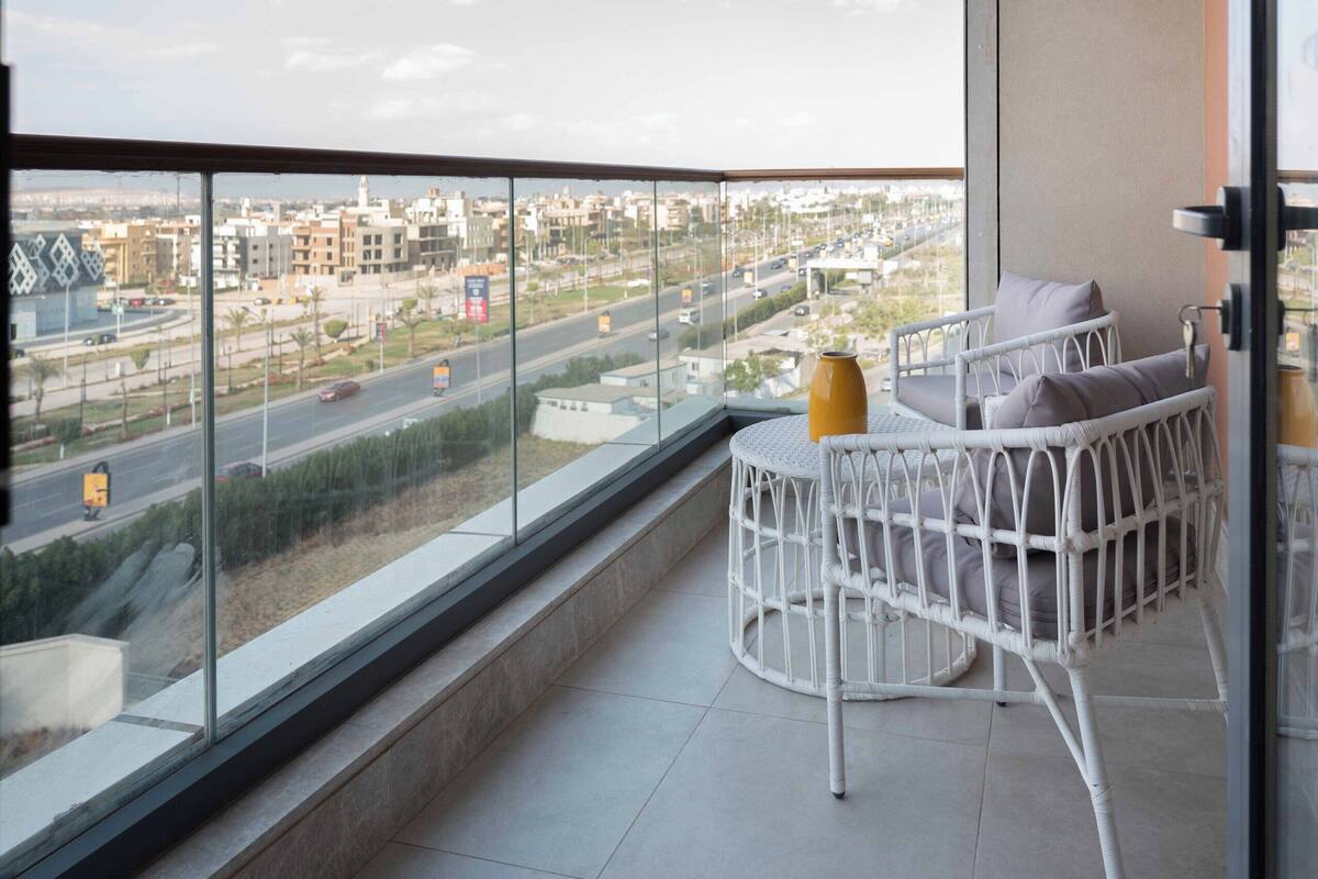 A balcony with a white wicker chair and a yellow vase, overlooking a cityscape with buildings and roads.