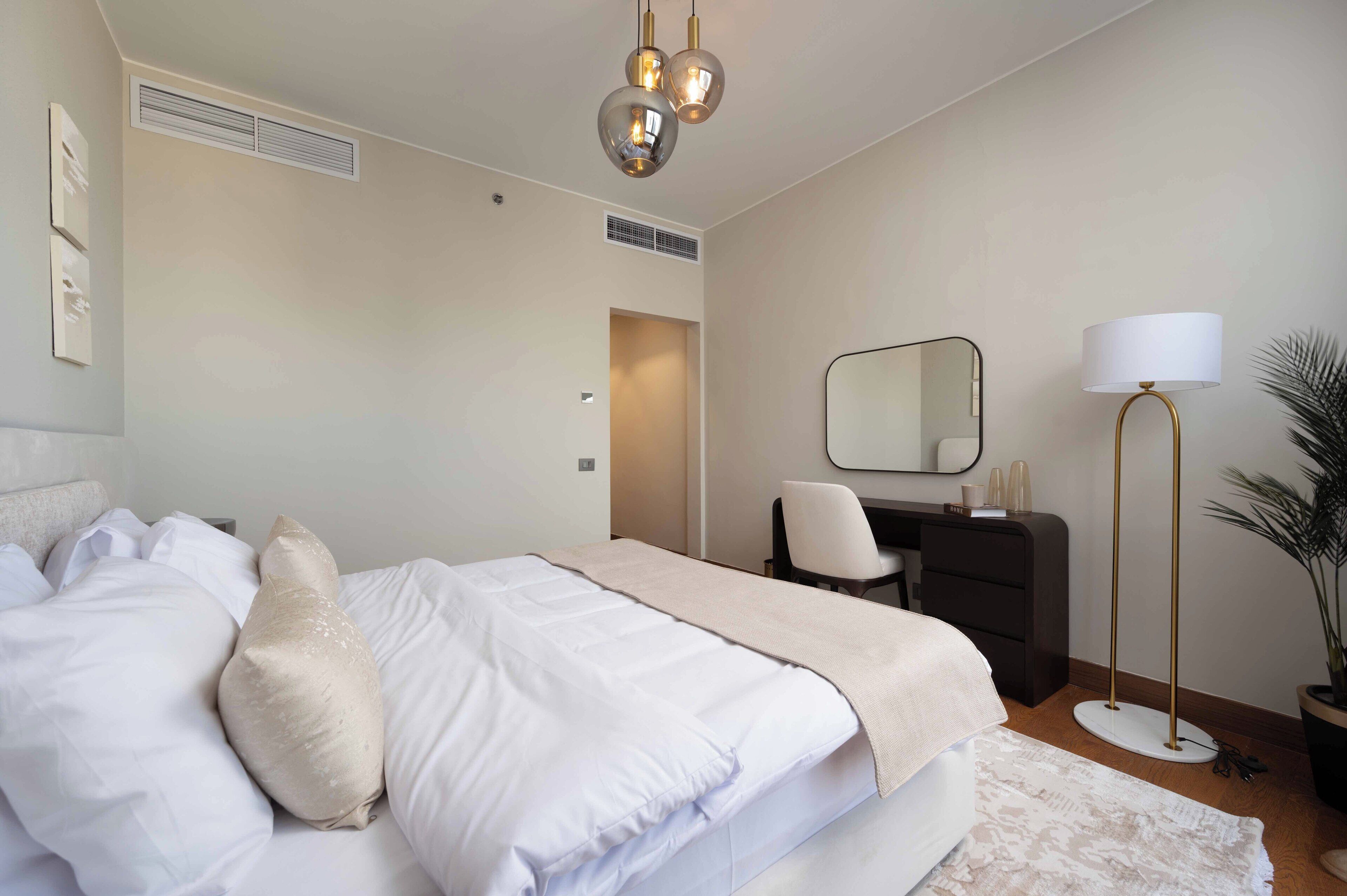 A neatly made bed with white linens and a beige throw blanket, a desk with a chair, a standing lamp, and a mirror on the wall.
