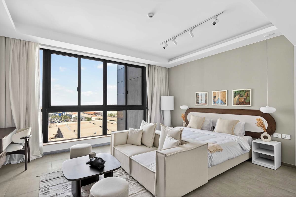 A modern hotel room with a large window, a sofa, a bed, a desk, and a chair.