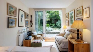 Una habitación acogedora con una cama, dos sillones, una mesita y una lámpara. Se observan cuadros en las paredes y vistas a zonas verdes al exterior.