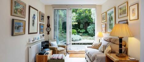 A cozy room with a bed, two armchairs, a small table, and a lamp. There are framed pictures on the walls and a view of greenery outside.