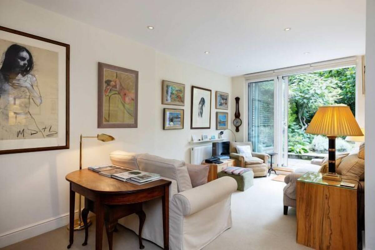 A modern living room with a sofa, a coffee table, and a lamp. There are framed artworks on the wall and a large window providing a view of greenery outside.