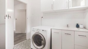 A modern laundry room with a front-loading washing machine, white cabinets, and a tiled floor. - Sammy's Seaside Hideaway in Henley Beach (Henley Beach South)