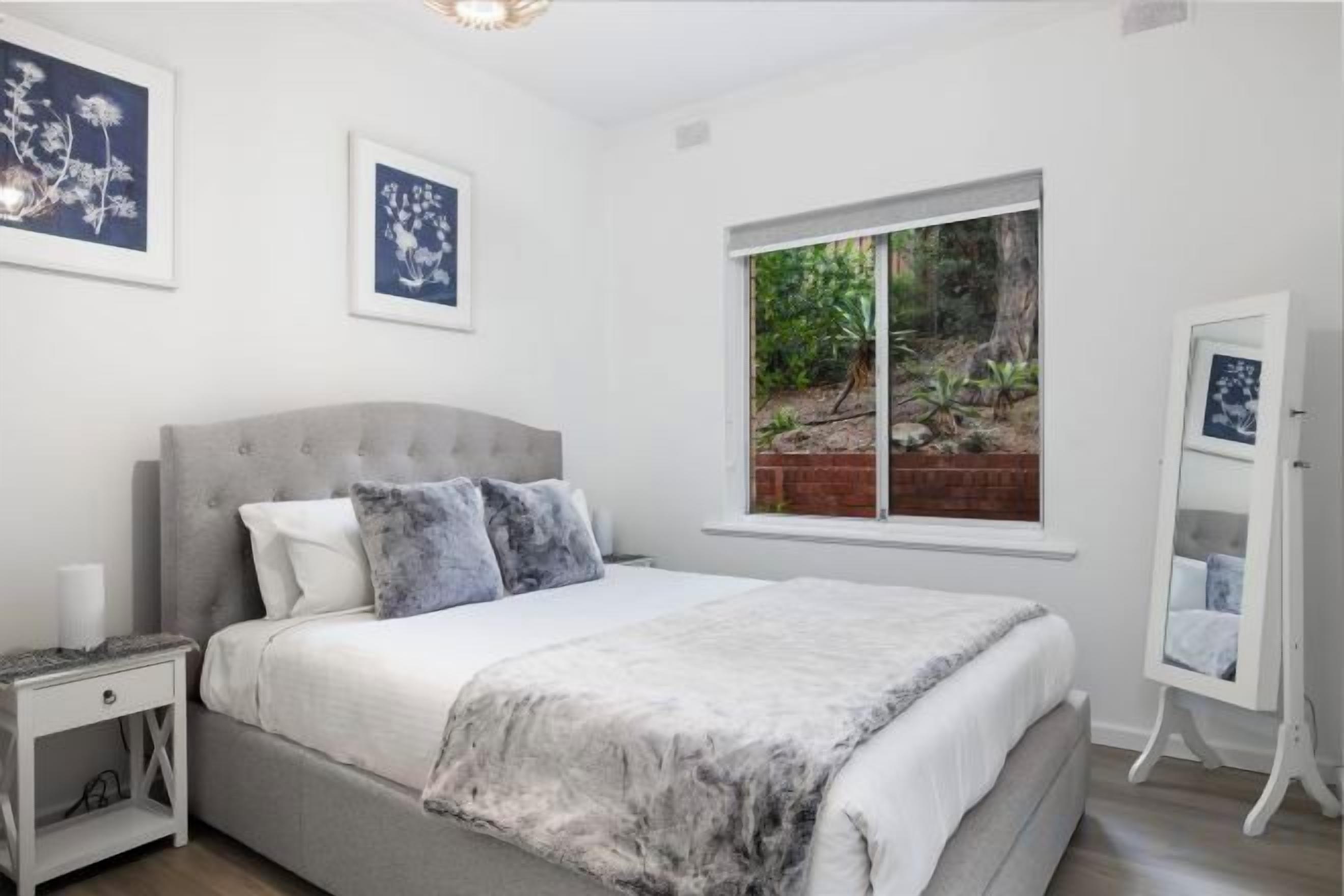 A bedroom with a bed, a nightstand, a window with a view of greenery, and a mirror on a stand.