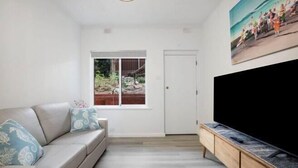 A living room with a sofa, a TV, and a window with a view of a garden. - Sammy's Seaside Hideaway in Henley Beach (Henley Beach South)