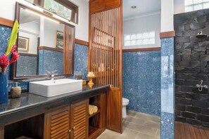 A bathroom with a large mirror, a white sink, a wooden cabinet, a shower with a stone wall, and a toilet. - Umah Bali Villa (Bali)