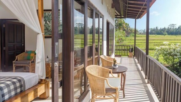 A bedroom with a bed, a chair, and a table on a balcony with a view of a green field. - Umah Bali Villa (Bali)