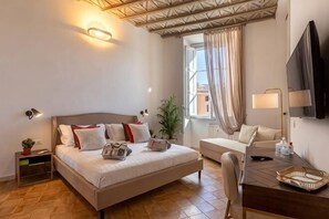 A hotel room with a bed, a desk, a chair, a TV, a window with curtains, and a plant. - Amelie Suite Navona (Rome)