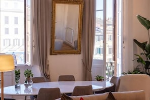 A hotel room with a round dining table, four chairs, a large mirror, and a view of buildings through large windows. - Amelie Suite Navona (Rome)