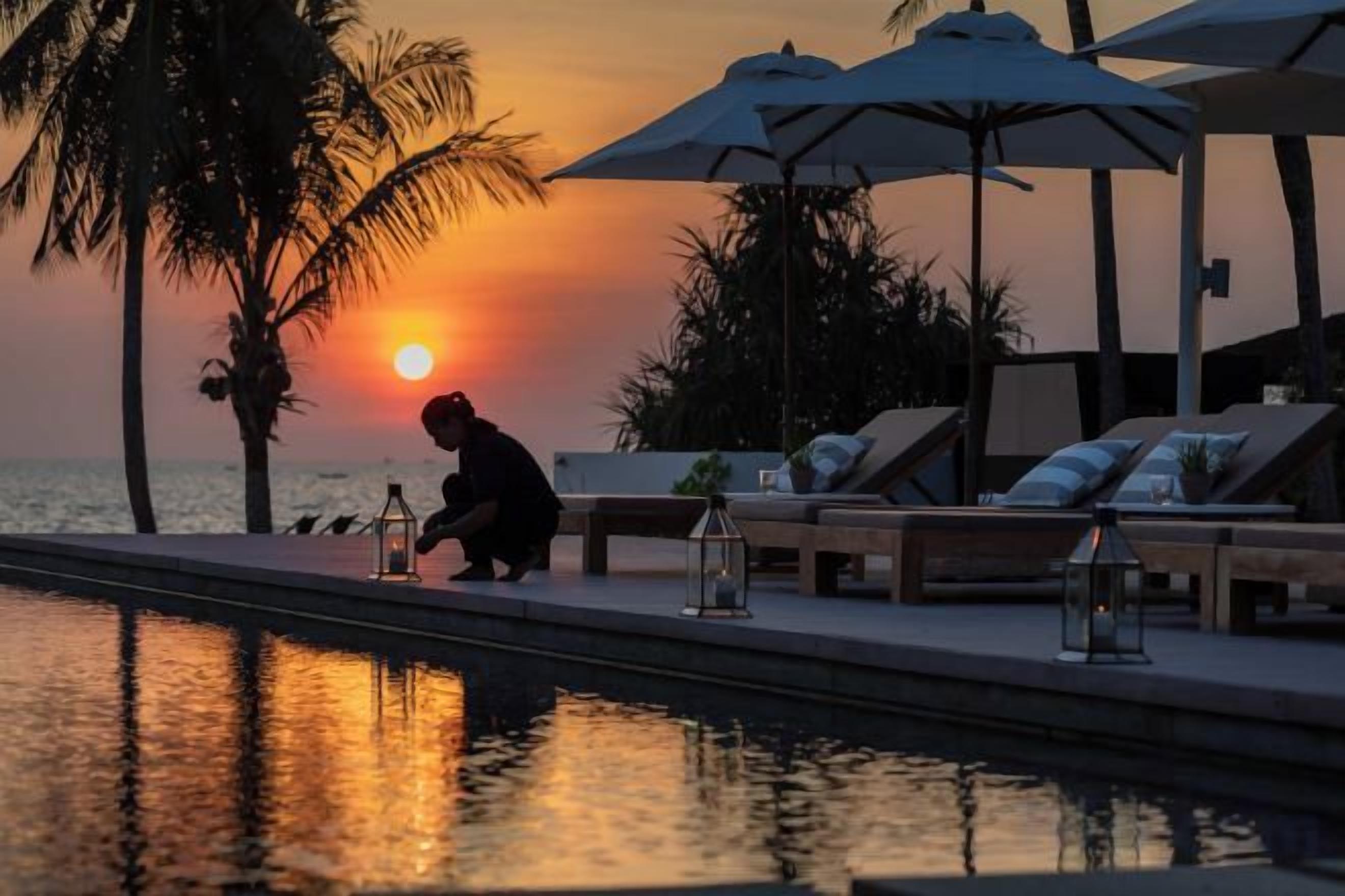 A poolside area with lounge chairs and umbrellas, a sunset in the background, and a person by the pool.