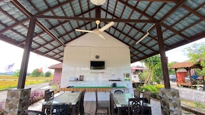 An outdoor covered dining area with a table and chairs, a ceiling fan, and a television mounted on the wall. - Capital O 91227 Chalet D'bendang Mak Teh (Pendang)