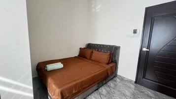 A single bed with a brown mattress and headboard, a small bedside table, and a dark door.