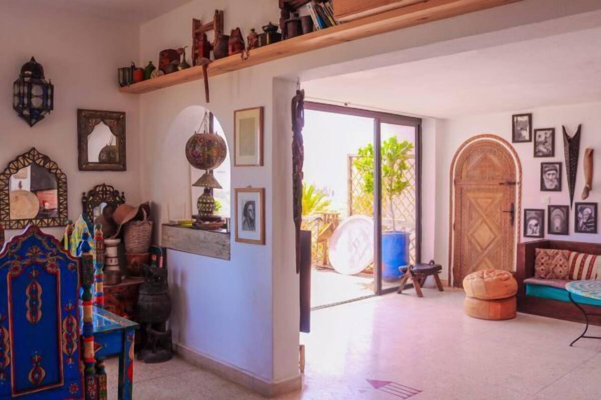 Une pièce avec un plafond en bois, un fauteuil bleu orné, une porte en bois et une vue sur l’extérieur.