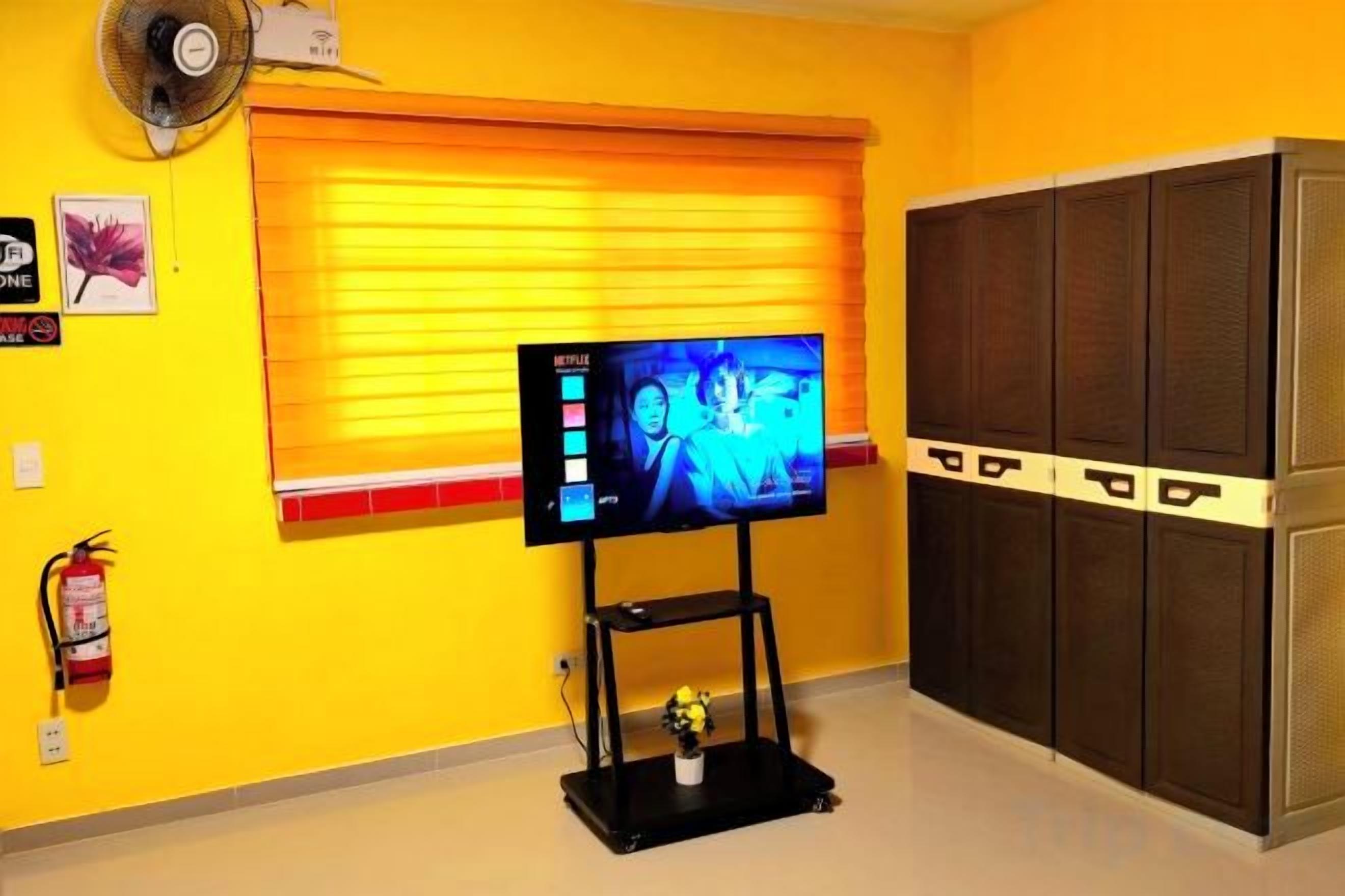 A room with a television on a stand, a window with blinds, a fire extinguisher, and a storage unit.