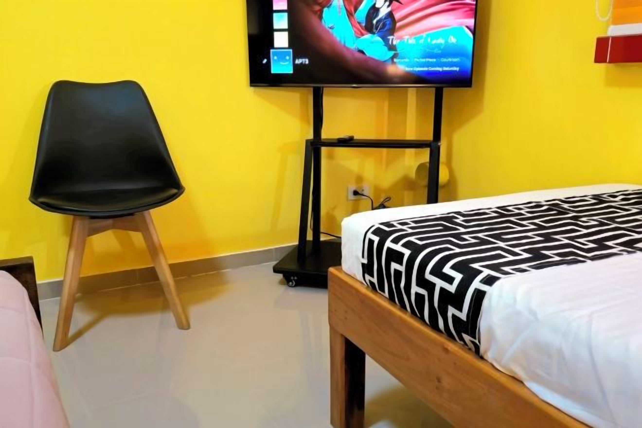Une chambre d’hôtel avec un lit, un téléviseur fixé au mur, une chaise noire et un mur jaune.