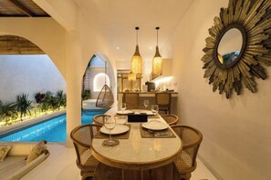 Un comedor con vista a la piscina, una mesa redonda con sillas, lámparas colgantes y un espejo con diseño de sol en la pared.