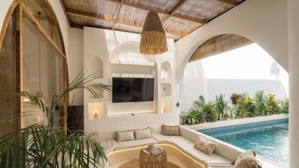 A modern living room with a curved sofa, a TV mounted on the wall, and a pool visible through an archway. - Brand New Villa 2Br with Private Pool Near Seminyak by BaliSuperHost (Bali)