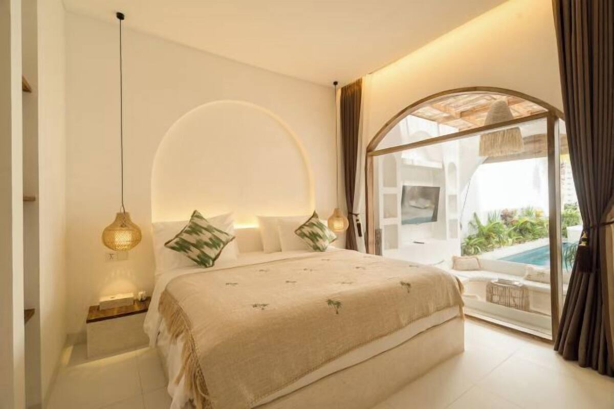 A bedroom with a bed, bedside tables, a hanging light fixture, and a view of an outdoor pool area.