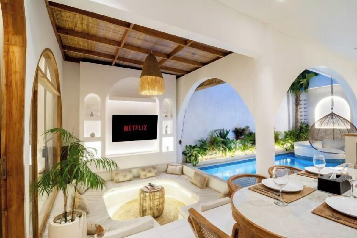 Un área interior de piscina moderna con zona de estar, una mesa de comedor para dos personas y una pantalla de televisión que muestra 'NETFLIX'.
