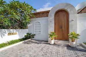 Entrada de una villa con puerta de madera, paredes blancas y plantas en macetas.