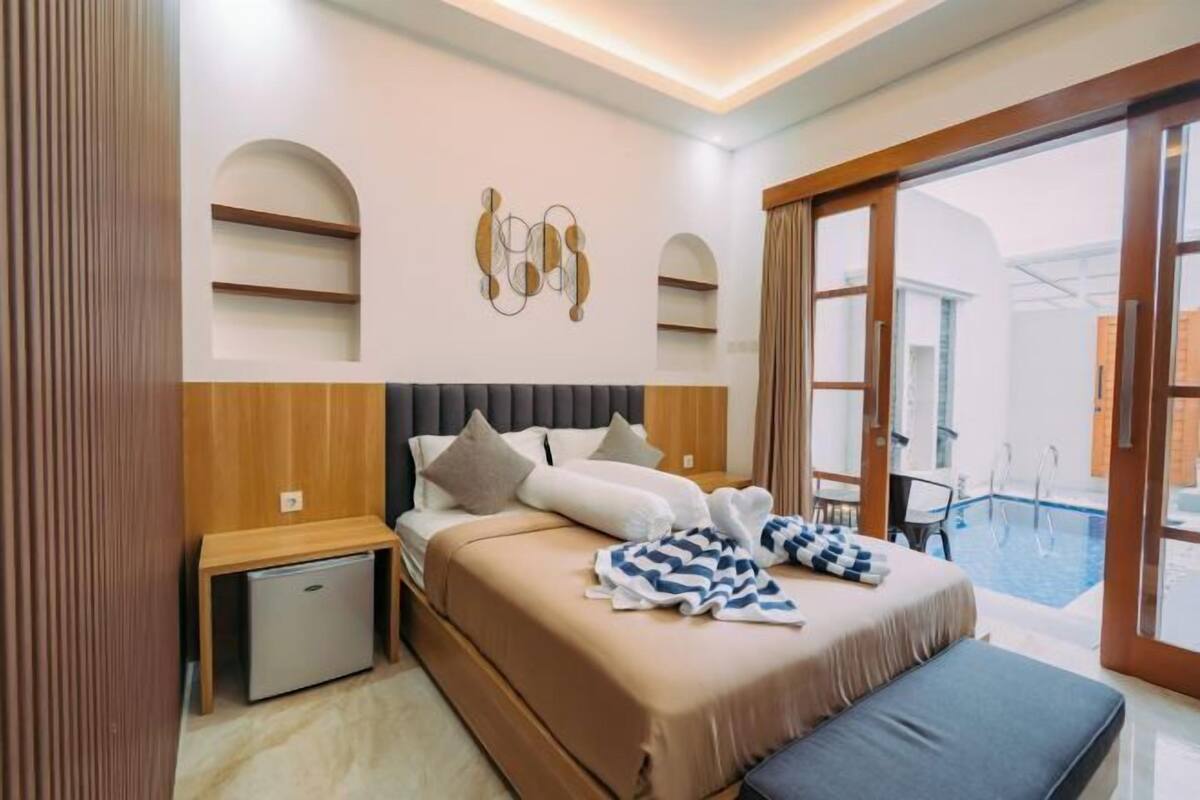 Una habitació d'hotel moderna amb un llit gran, un escriptori petit, un frigorífic i vista a una piscina exterior.