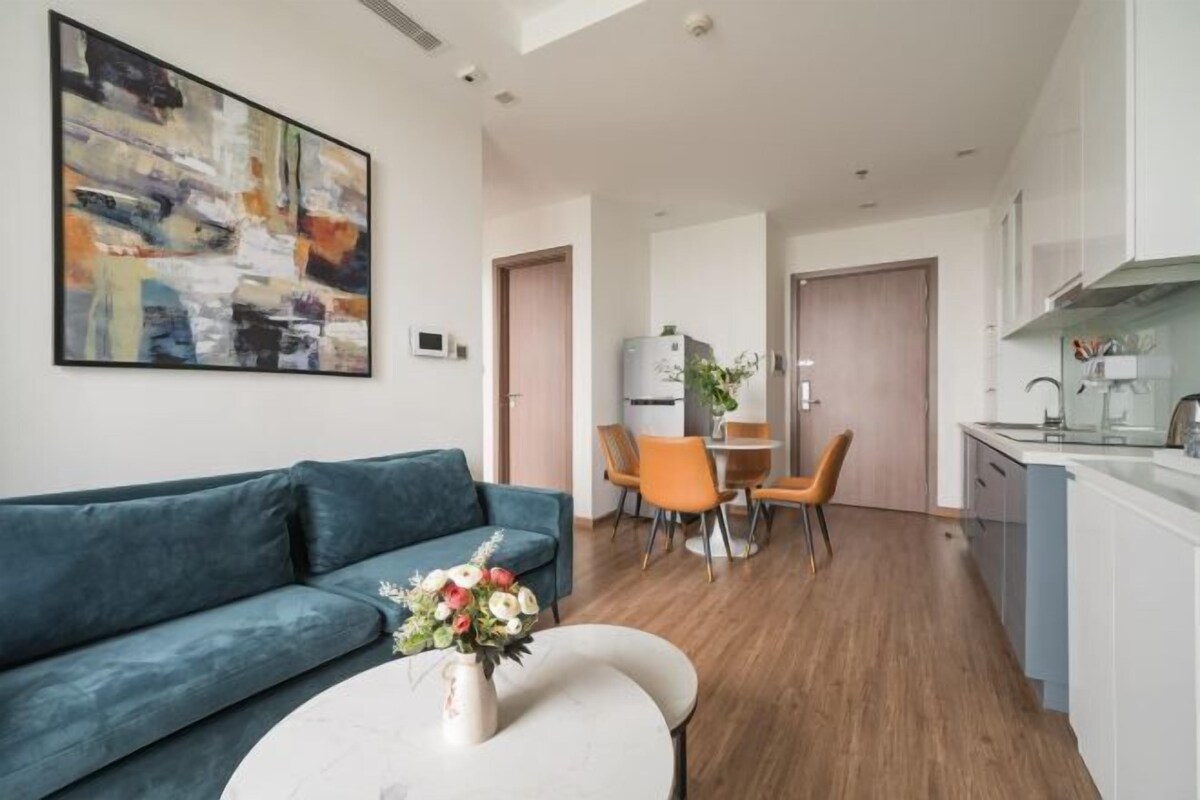 A modern living room with a sofa, a dining area with chairs, a kitchen with cabinets, and a large abstract painting on the wall.