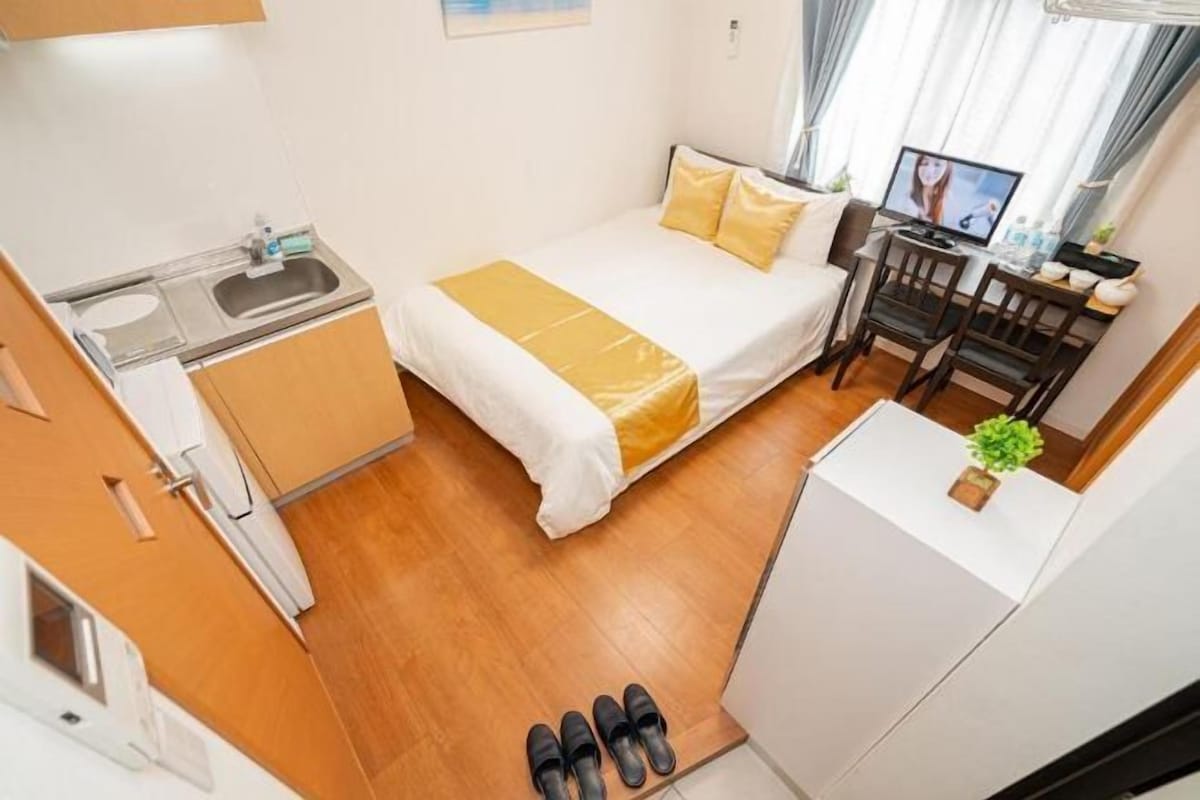 A compact hotel room with a bed, a desk with a computer, a small kitchenette, and a pair of slippers on the floor.