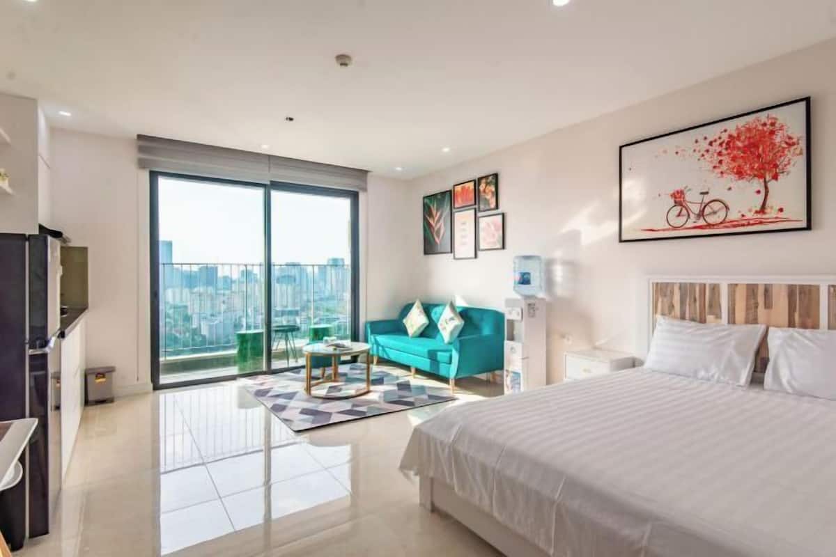 A modern hotel room with a large bed, a sofa, a dining table, and a city view through the balcony.