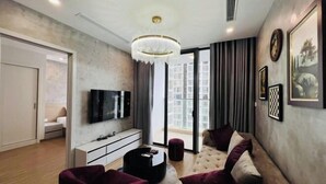 A modern living room with a flat-screen TV, a chandelier, a sofa, and a large window with a city view. - Alex'housing Vinhome D'capitale luxury apartment (Hanoi)