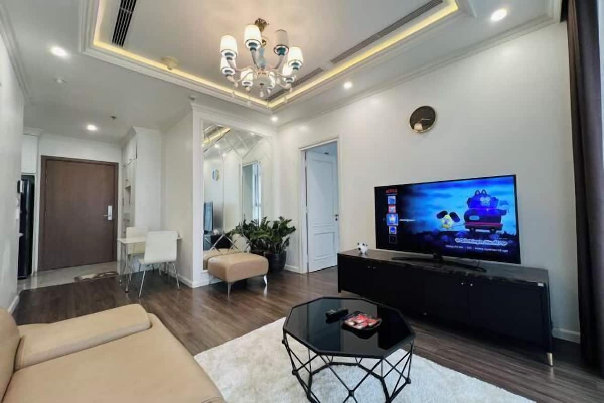 A modern living room with a sofa, a glass coffee table, a TV on a black stand, and a chandelier.