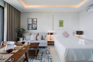 A modern hotel room with a bed, a sofa, a dining table with wine glasses, and a lamp.