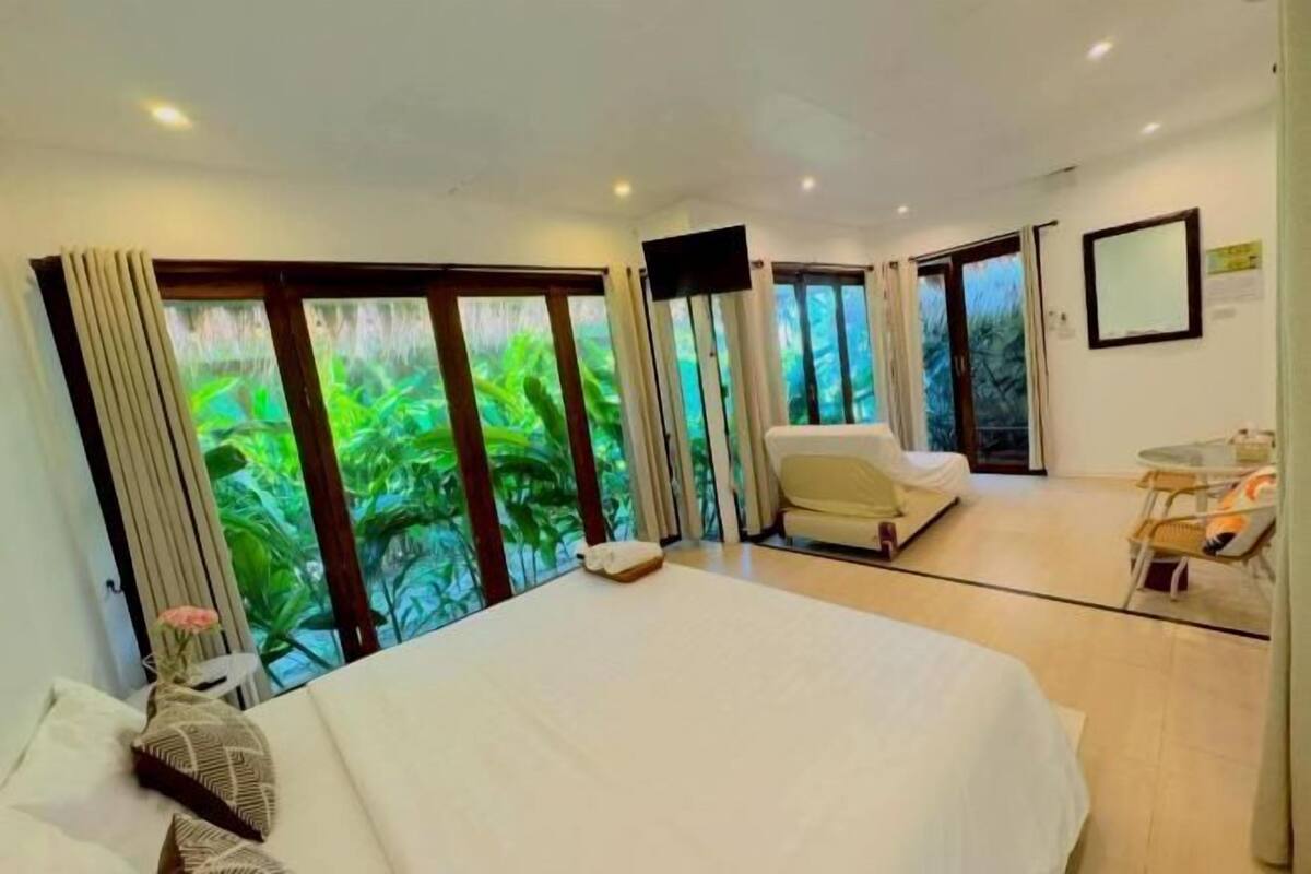 A hotel room with a bed, a chair, a small table, and large windows with a view of greenery.
