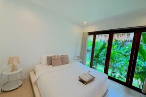 Een slaapkamer met een groot bed, een nachtkastje met een lamp en uitzicht op groen door een raam.