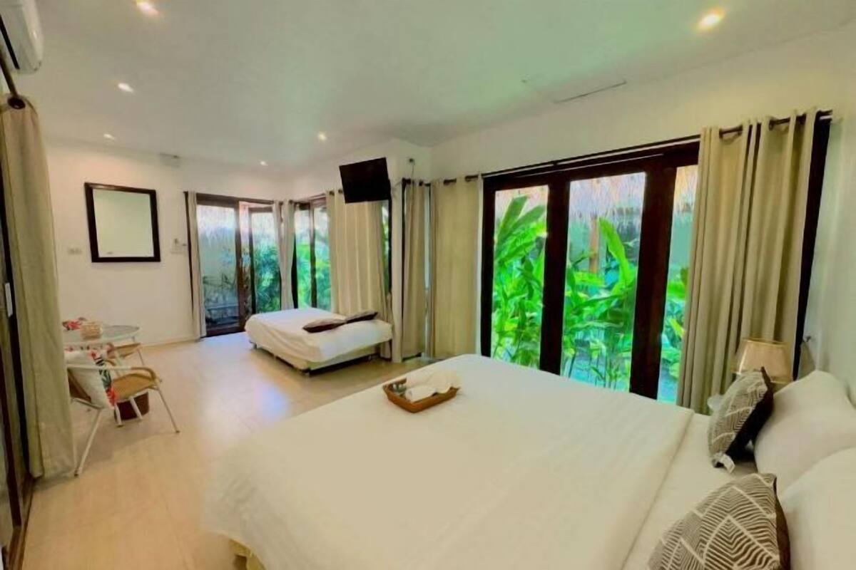 A hotel room with a large bed, a desk with a chair, and a view of greenery through the windows.