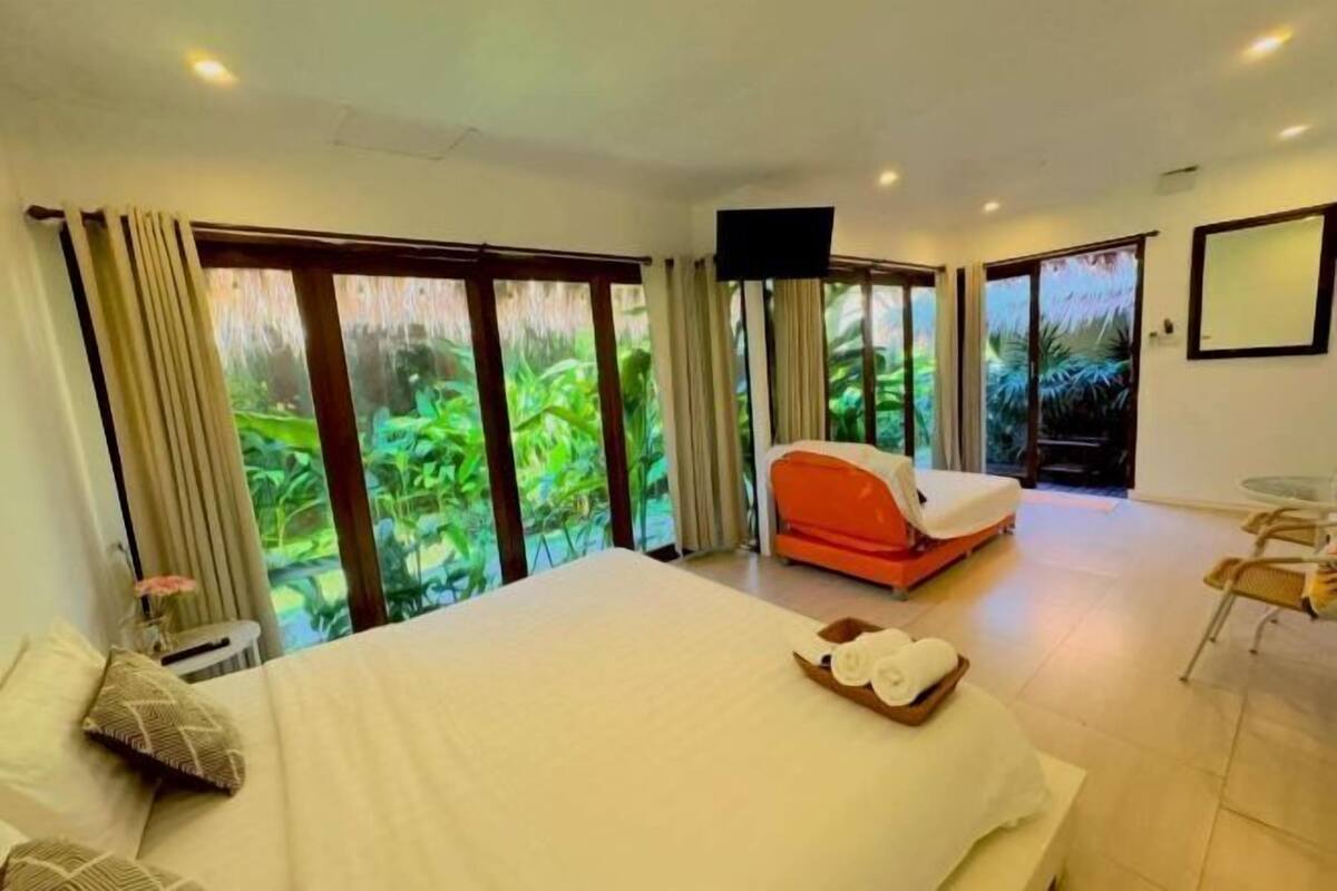 A hotel room with a bed, a massage chair, a TV, and large sliding glass doors to a garden.