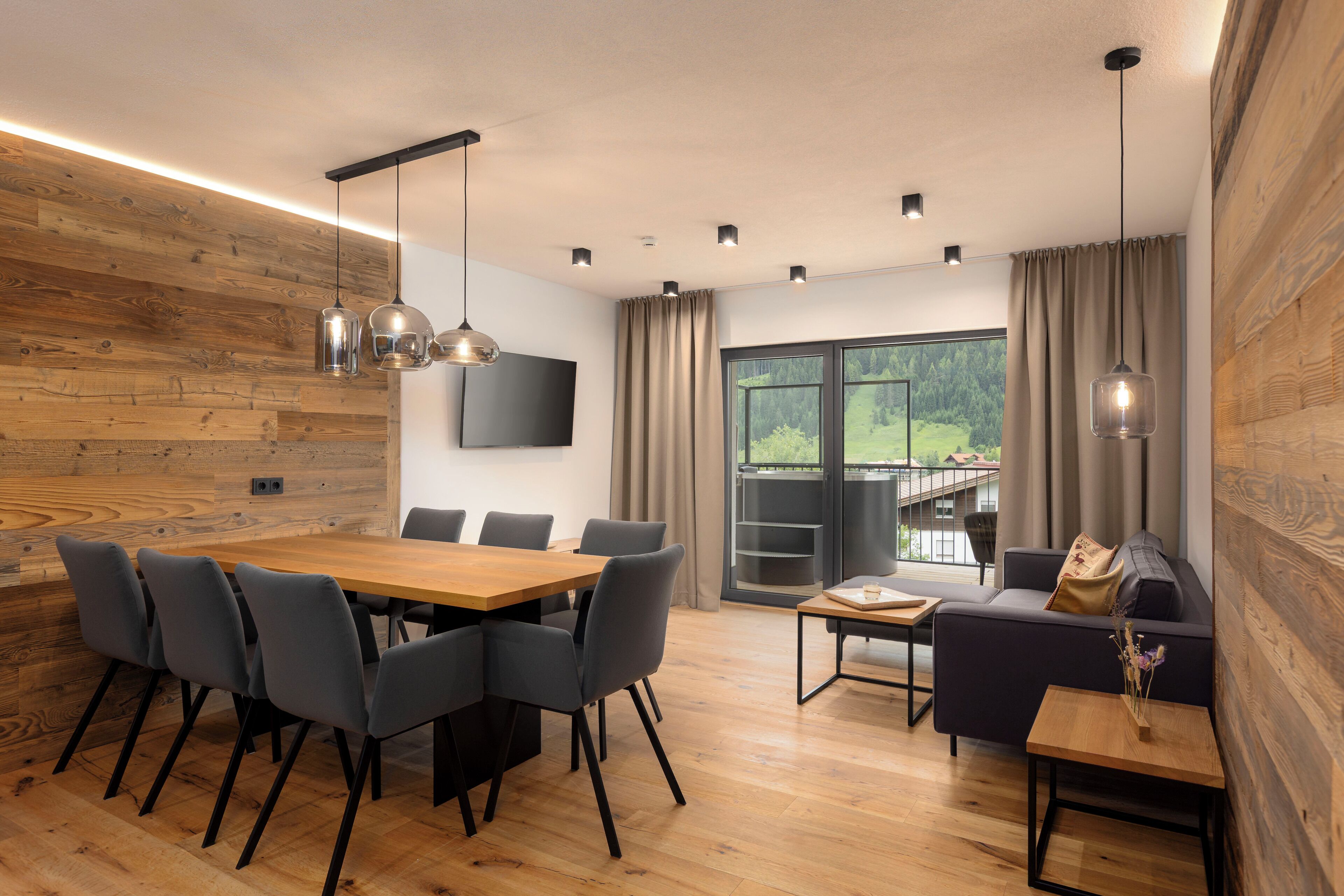 Un intérieur moderne avec un mur en bois, un coin repas comprenant une table et des chaises, un canapé et une grande fenêtre donnant sur un paysage verdoyant.