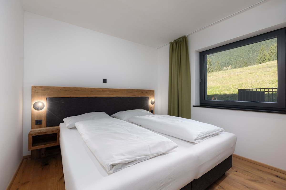 A bedroom with a wooden headboard, two beds, a nightstand, and a window with a view of a green landscape.