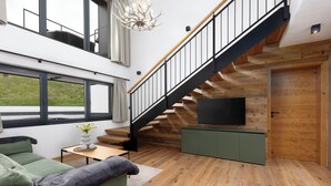 A modern living room with a staircase, wooden flooring, a green sofa, a small table with a vase, and a flat-screen TV mounted on a wooden wall. - Bergresort Berwanger Hof by ALPS RESORTS (Berwang)