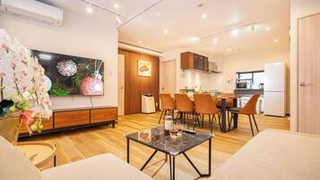 A modern living room with a flat-screen TV, a dining area with chairs, and a kitchen in the background.