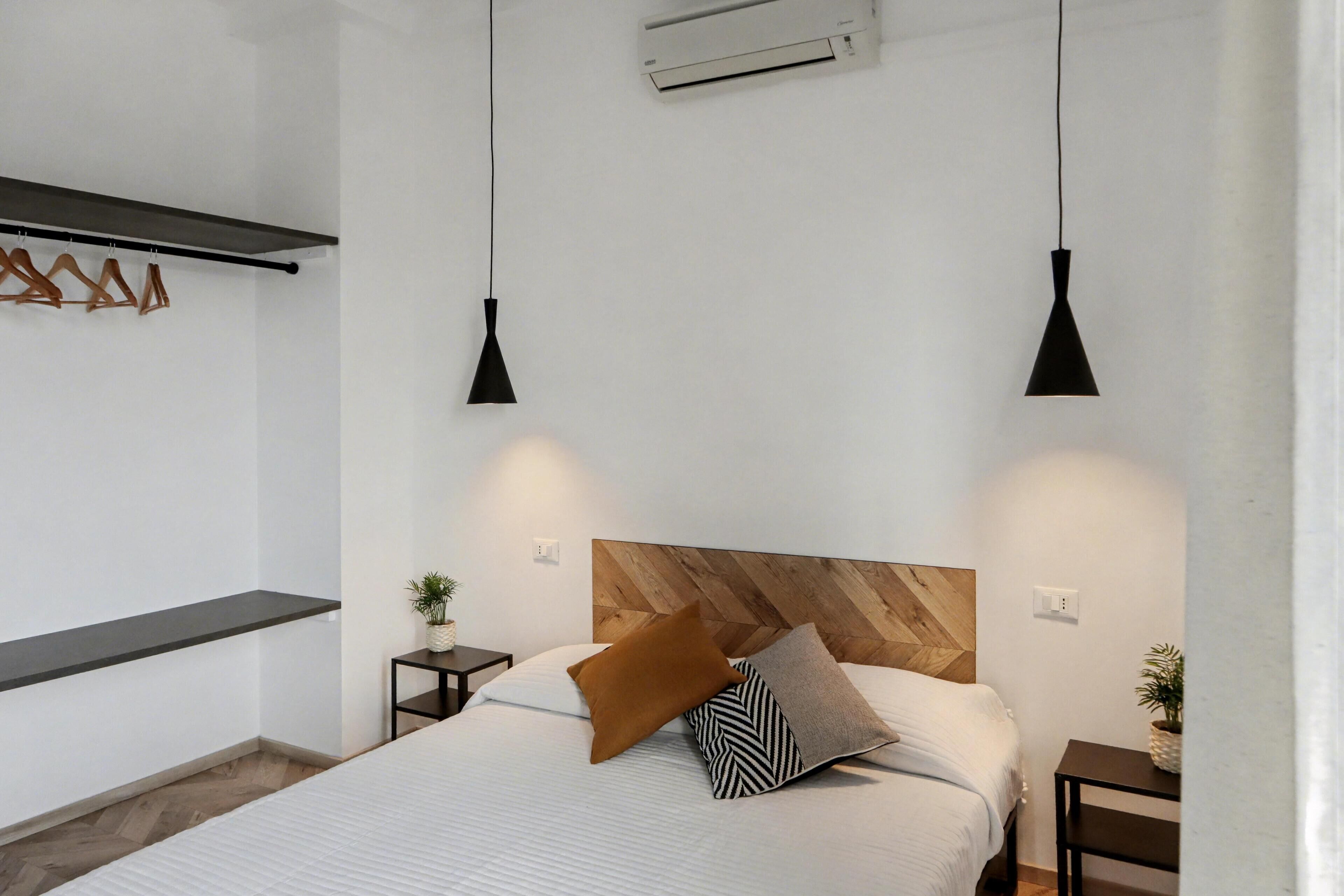 Un dormitorio moderno con cabecera de madera, cama con ropa de cama blanca, dos lámparas colgantes negras, una mesita de noche con una planta y una estantería de madera.