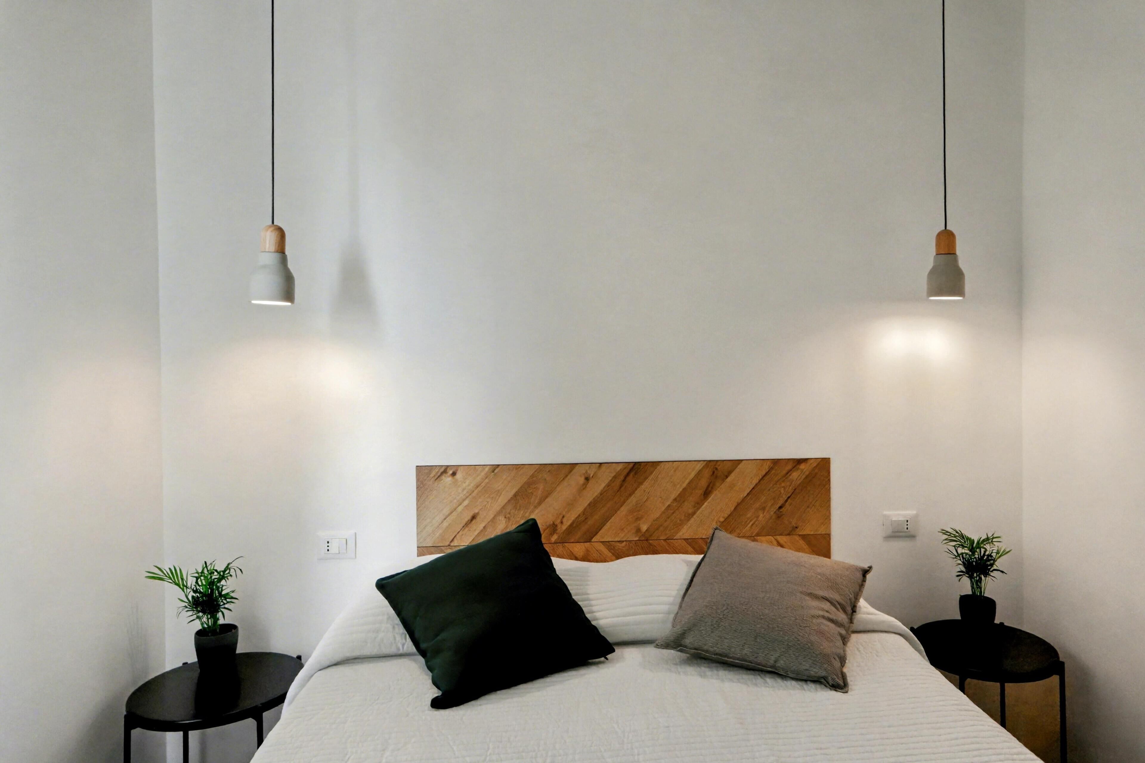 A modern bedroom with a wooden headboard, a bed with white bedding, two bedside tables with potted plants, and pendant lights.