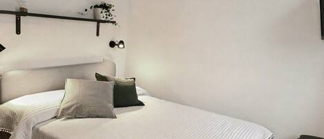 A neatly made bed with a white bedspread and pillows, a bedside table with a lamp, and a shelf with a plant and glasses.