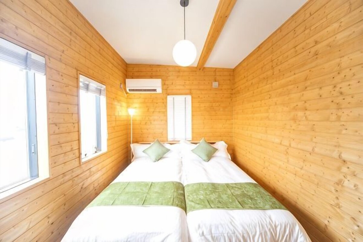 Una habitación con paredes de madera, una cama con almohadas verdes, una lámpara de noche y una ventana.