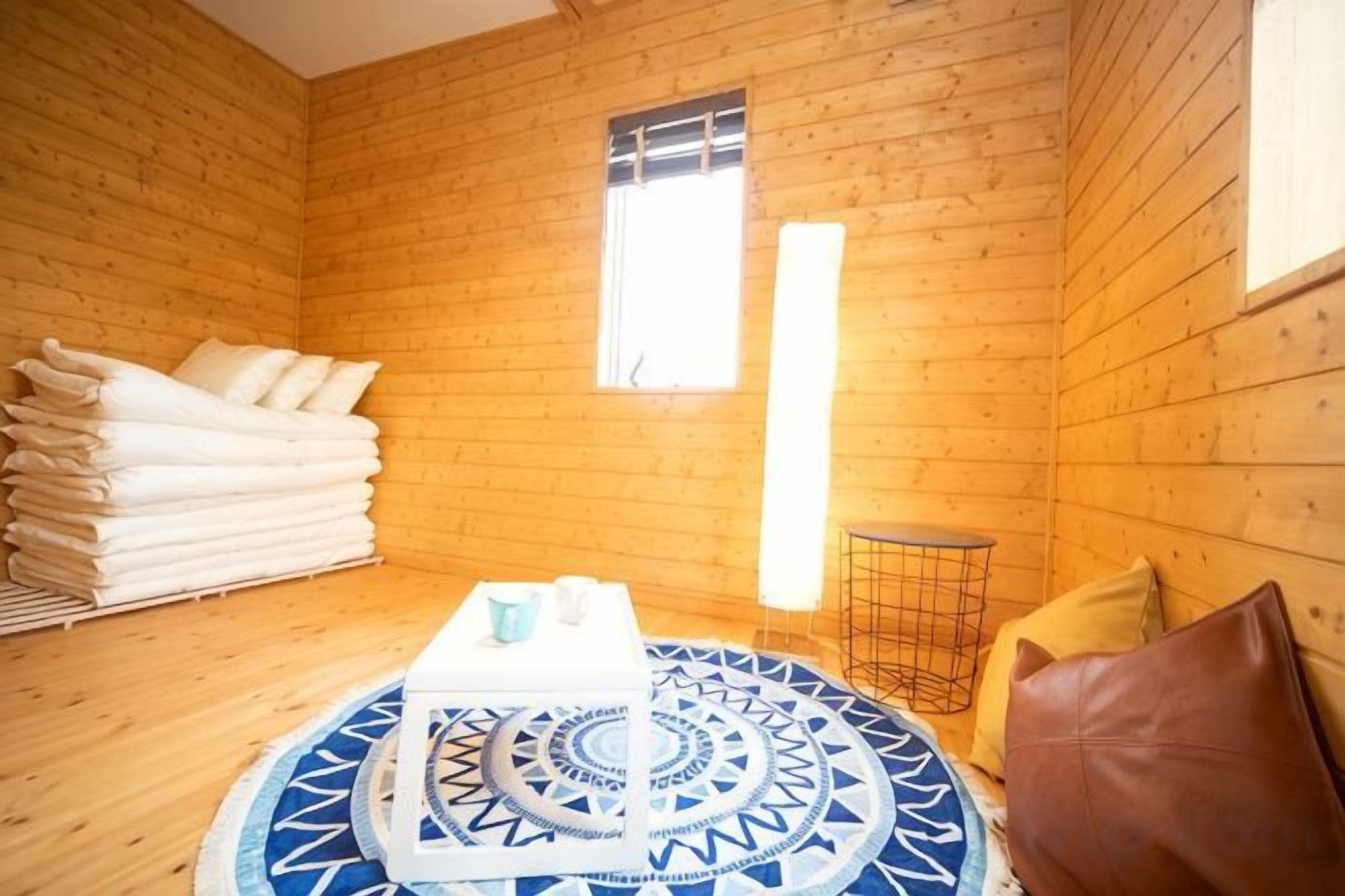 Una habitación con paredes de madera, una cama con ropa blanca, un tapete azul redondo con diseño, una pequeña mesa con un objeto blanco, y una ventana con persianas.